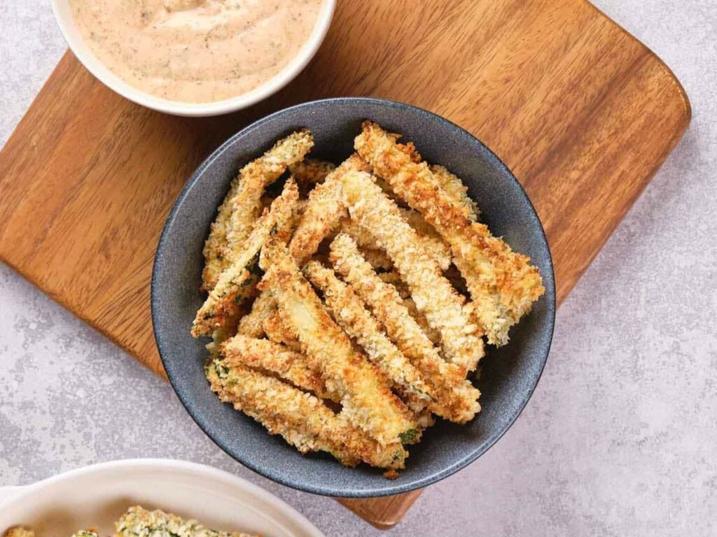A bowl of crispy, breaded zucchini fries sits on a wooden board.