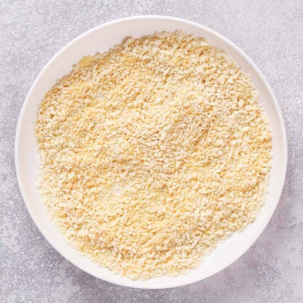 A white bowl filled with pale yellow, dry panko breadcrumbs.