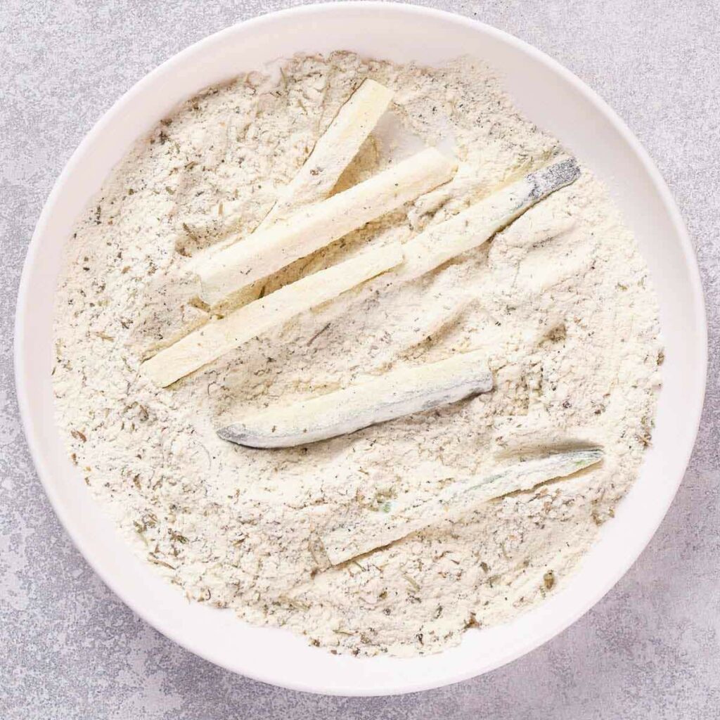 A white bowl containing flour and seasonings with several raw potato strips.
