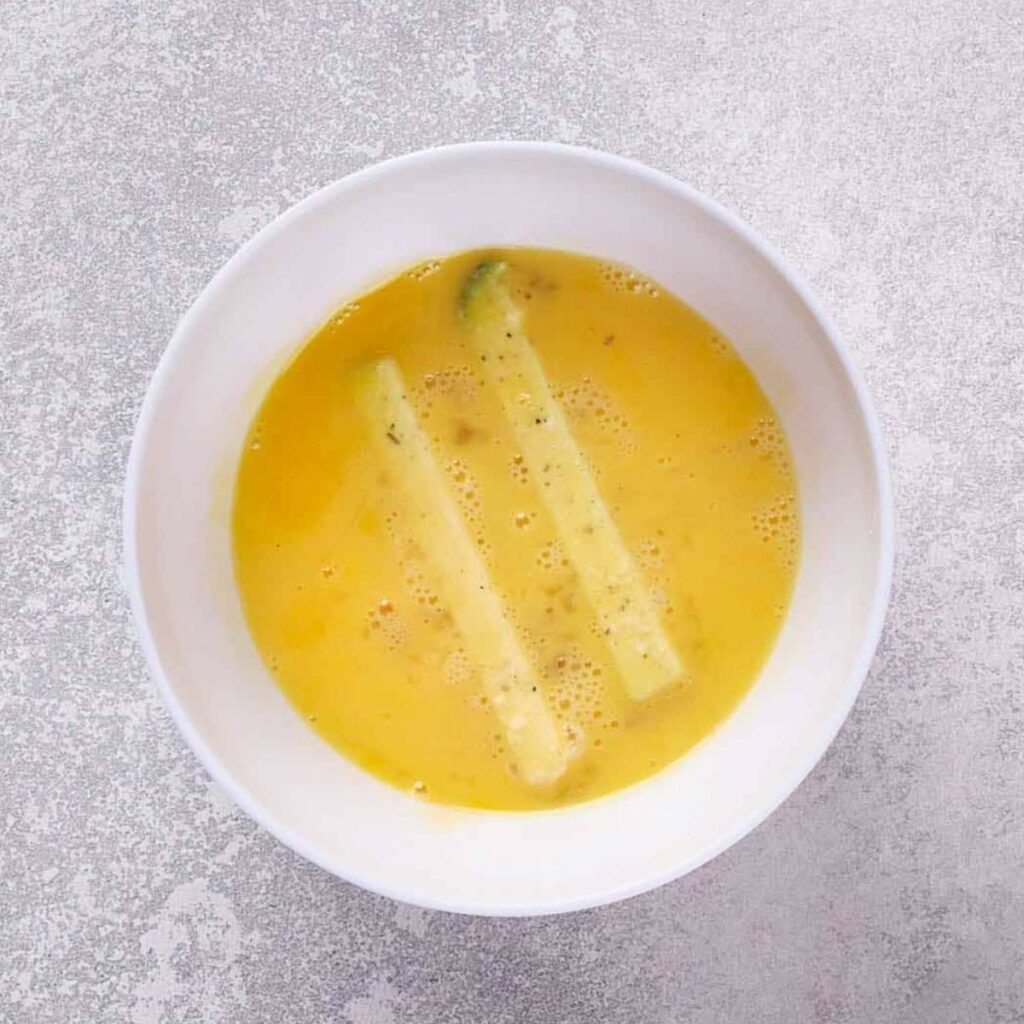 A white bowl containing whisked eggs with two pieces of zucchini strips.