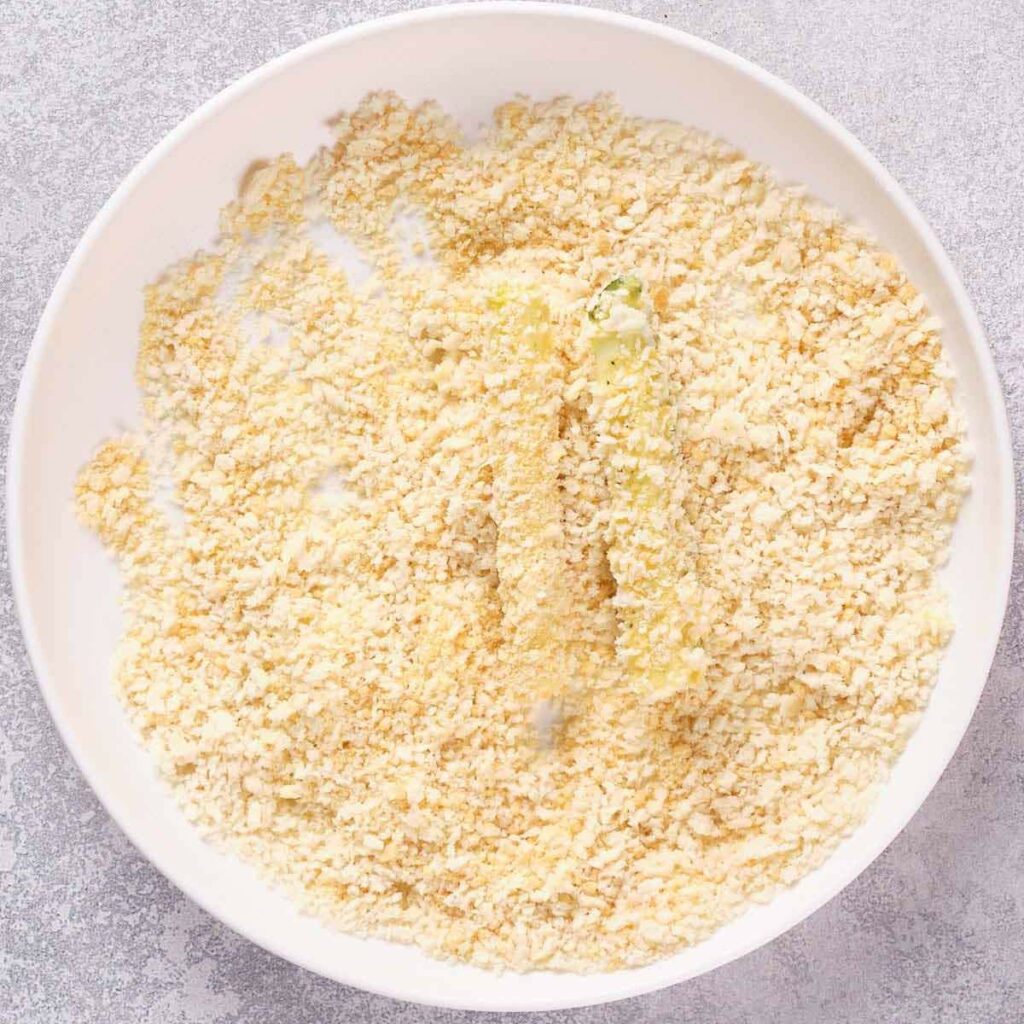 A white bowl containing a breadcrumb mixture with a coated zucchini strips.