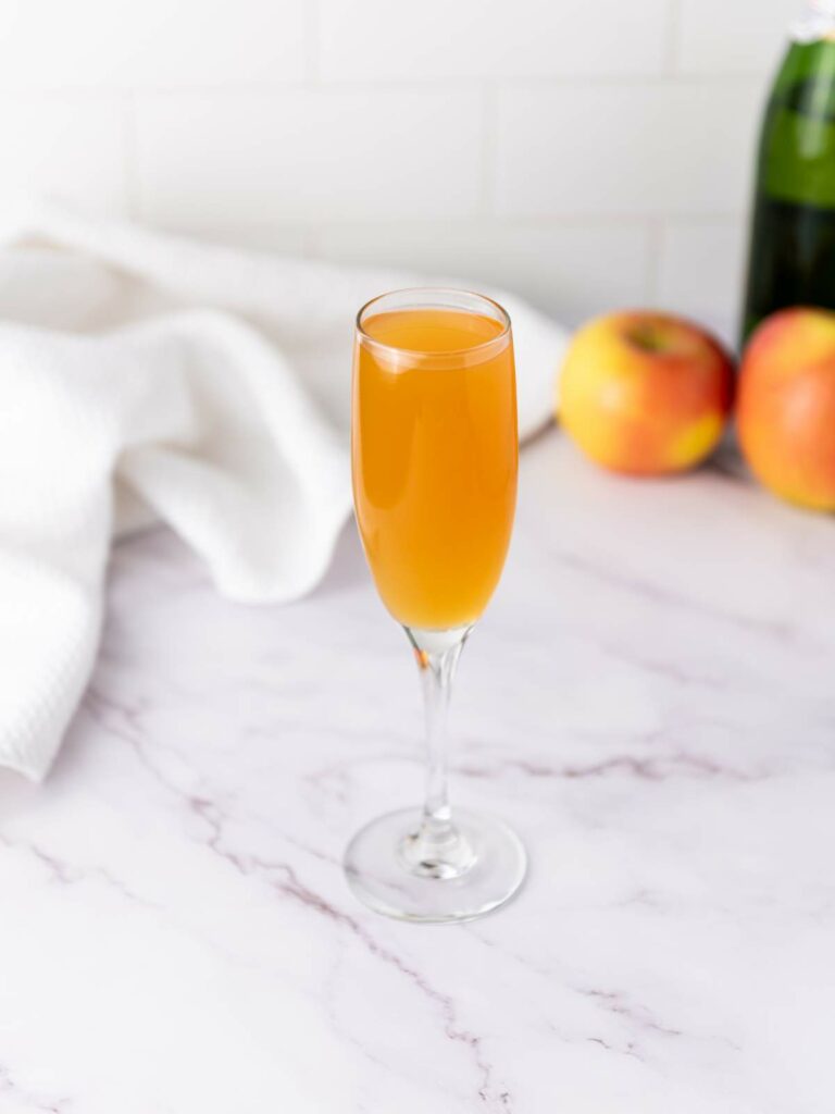 A champagne flute filled with an orange drink sits on a marble surface, with apples, a bottle, and a white cloth in the background.