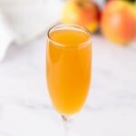 A champagne flute filled with mimosa sits on a white surface, with two apples blurred in the background.