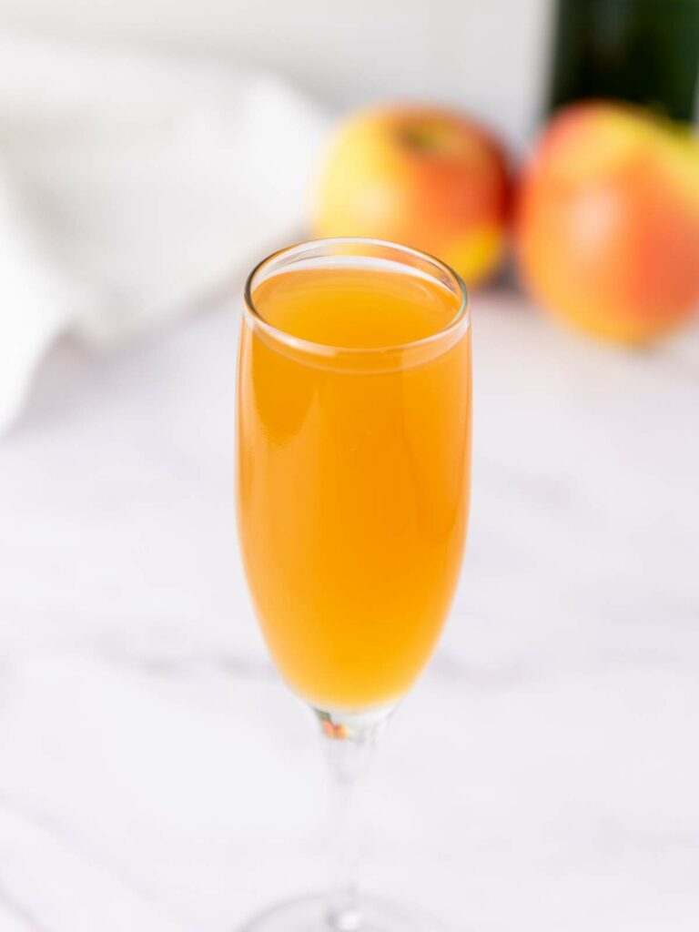 A champagne flute filled with mimosa sits on a white surface, with two apples blurred in the background.