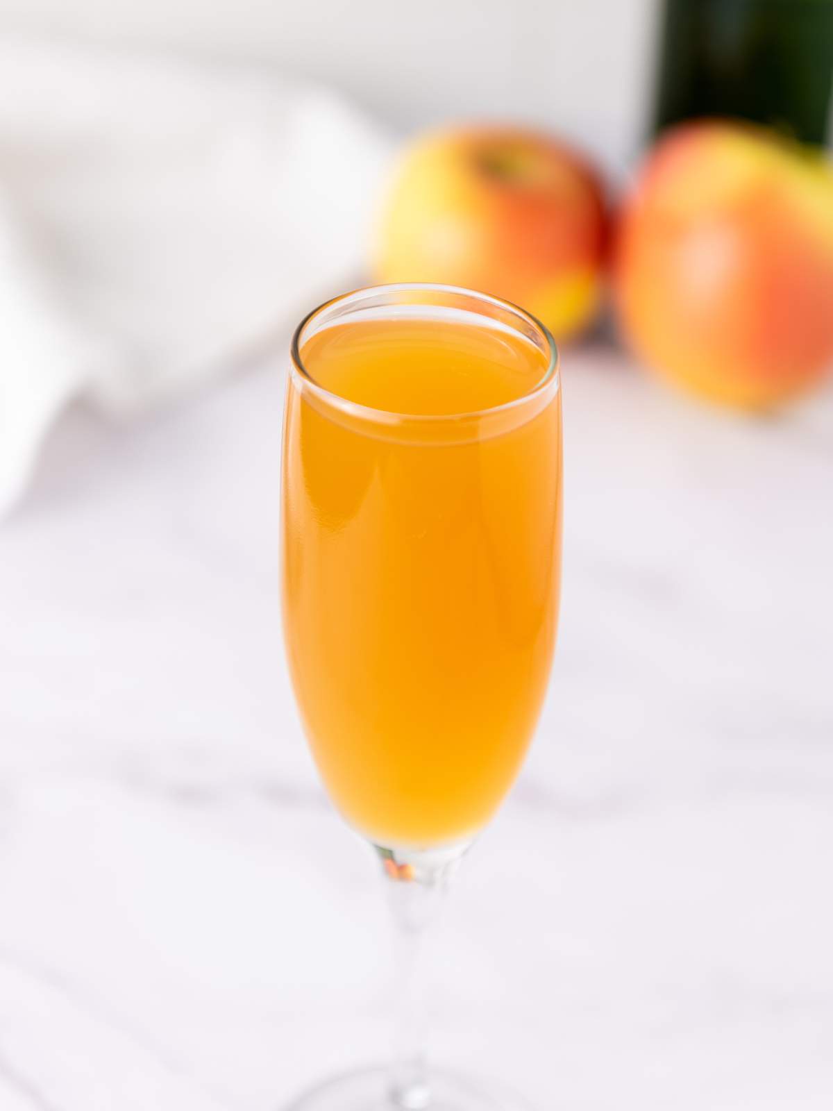 A champagne flute filled with mimosa sits on a white surface, with two apples blurred in the background.