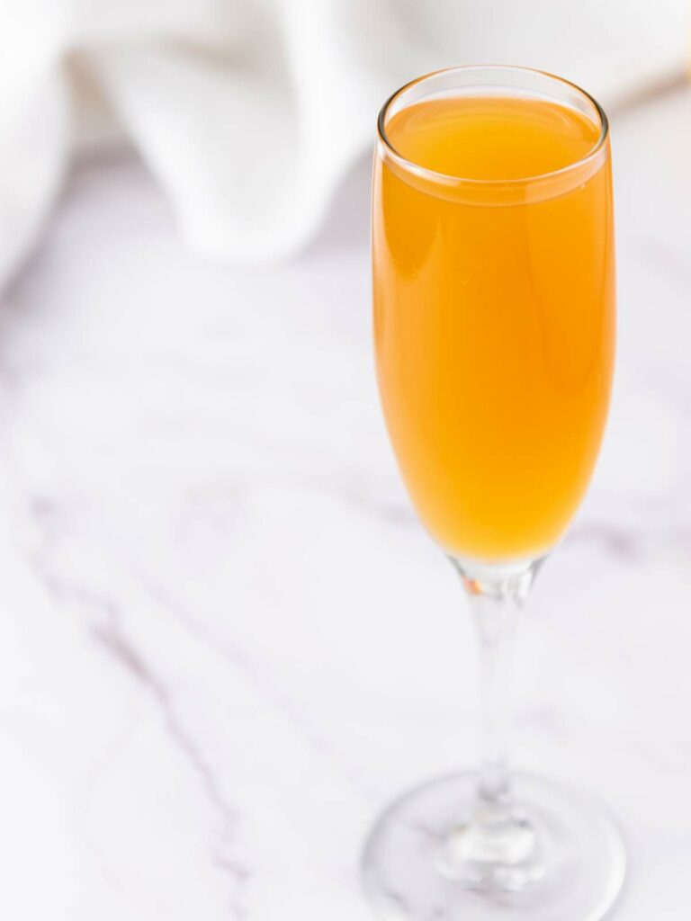 A champagne flute filled with orange-colored mimosa on a white marble surface.