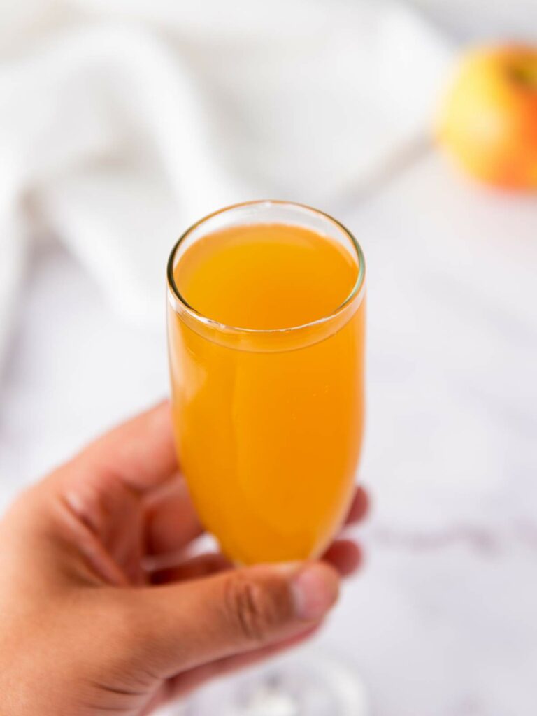 A hand holding a glass of orange-colored juice, with a white cloth and a blurred apple in the background.