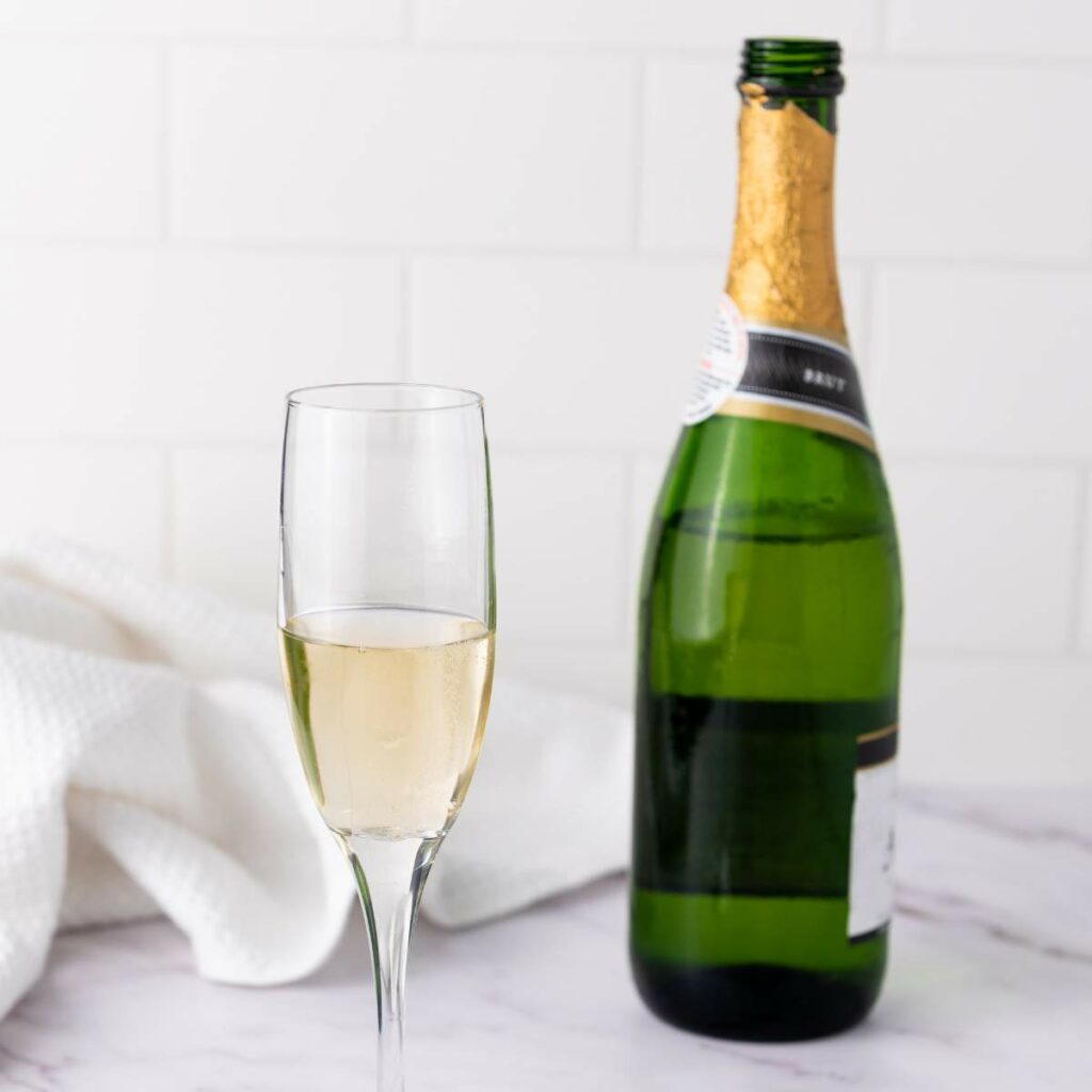 A glass of champagne sits next to an opened bottle on a marble surface with a white cloth in the background.