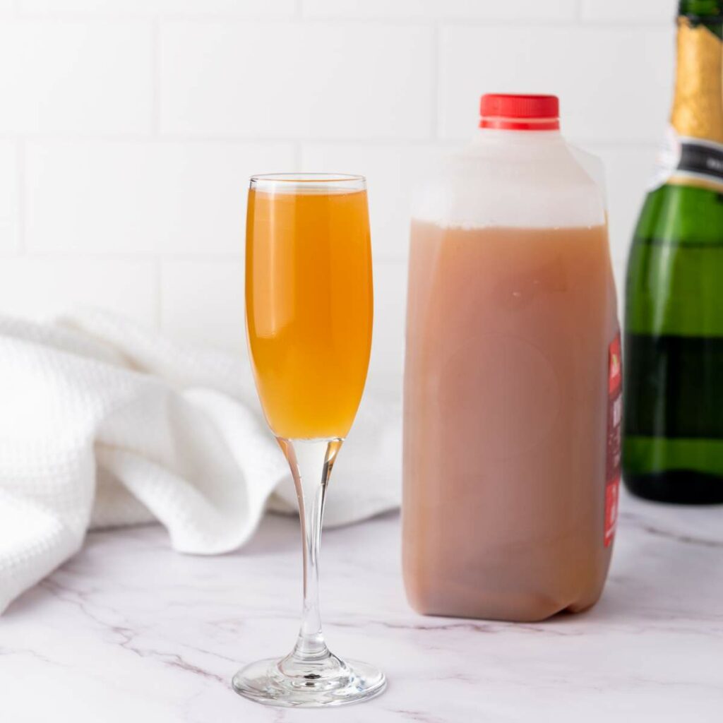 A flute glass filled with mixed champagne and apple cider sits next to a plastic jug of apple cider and a champagne bottle on a marble surface.