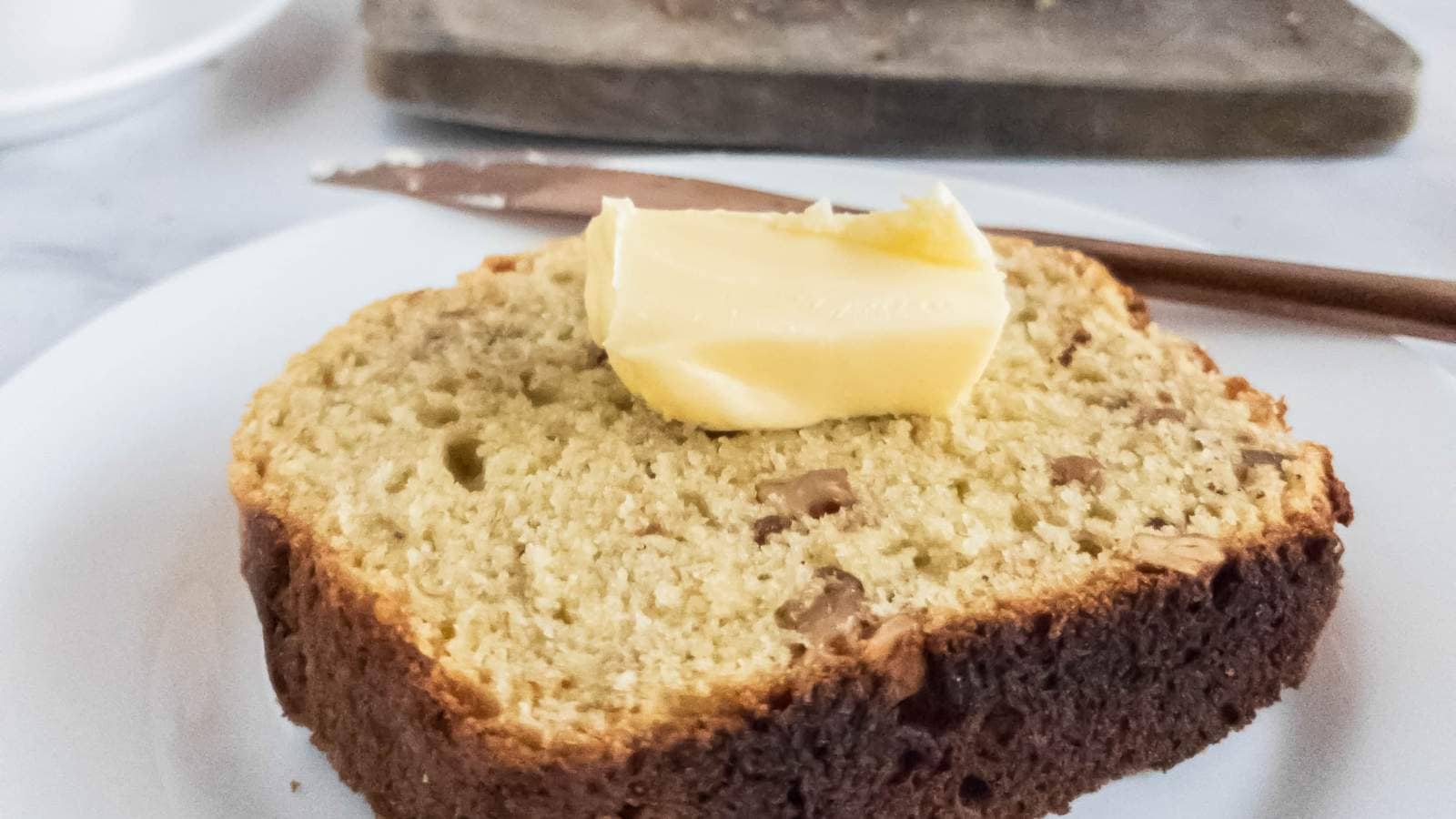 A thick slice of banana bread on a white plate with a dollop of butter on top.