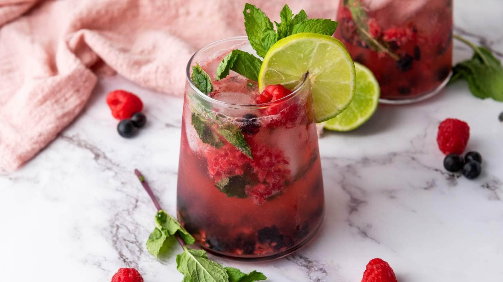 A glass of berry drink with crushed ice, fresh mint, lime slices, and mixed berries on a marble surface, with a pink cloth in the background.