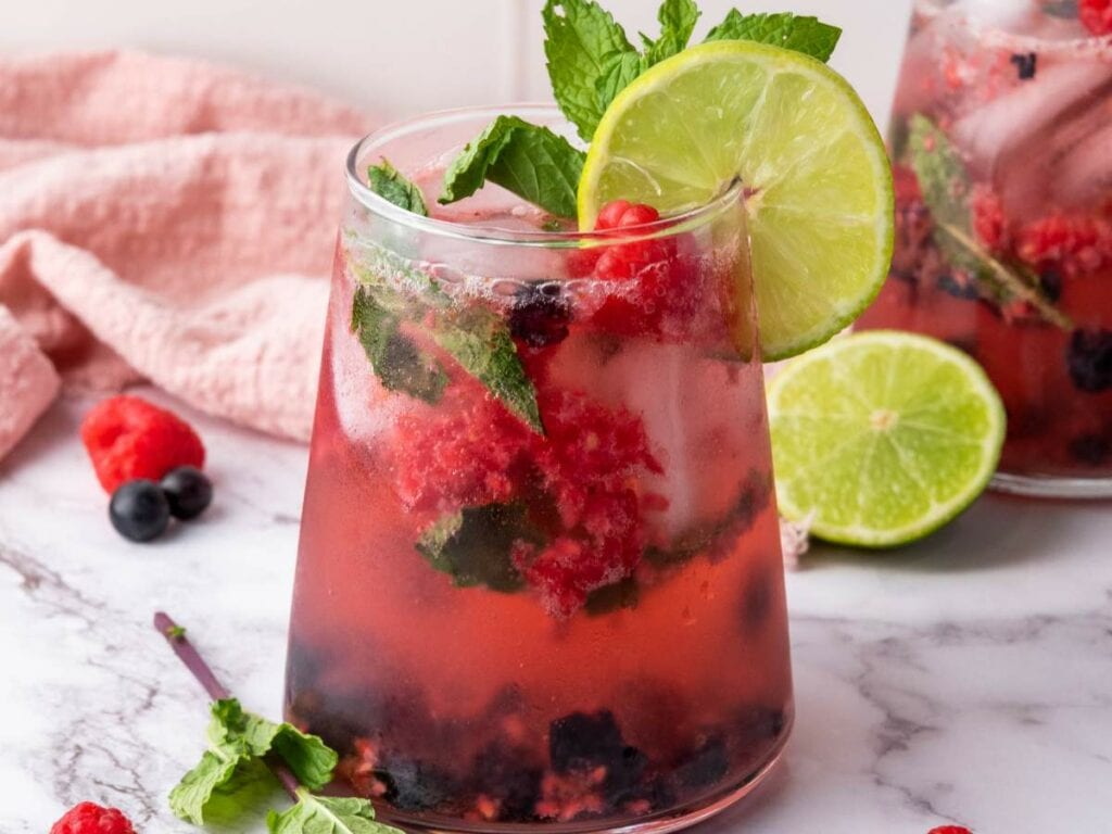 A glass of berry soda water with ice, mint leaves, crushed berries, and a lime slice garnish on a marble surface.