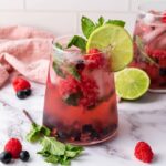 A glass of berry-infused drink with ice, lime slice, mint leaves, and mixed berries, set on a marble surface with a pink cloth and lime in the background.