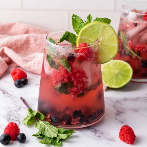 A glass of berry-infused drink with ice, lime slice, mint leaves, and mixed berries, set on a marble surface with a pink cloth and lime in the background.