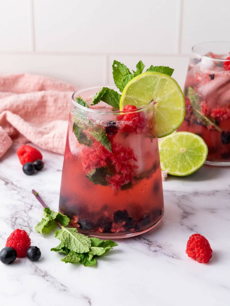 A glass of berry-infused drink with ice, lime slice, mint leaves, and mixed berries, set on a marble surface with a pink cloth and lime in the background.