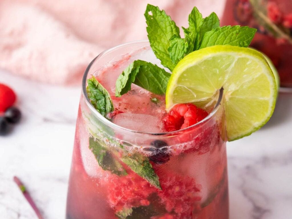 A glass of soda water with berries, mint leaves, and a lime slice garnish on a marble surface.