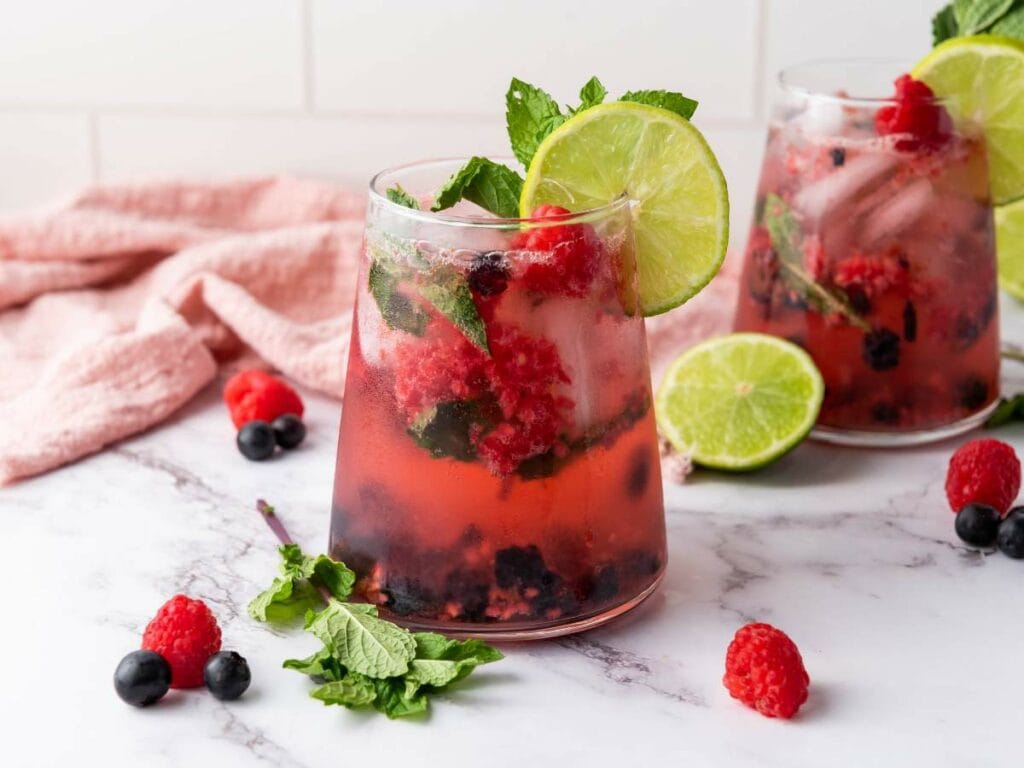 Two glasses of soda water garnished with lime slices, mint leaves, and mixed berries on a marble surface, with a pink cloth and scattered ingredients in the background.