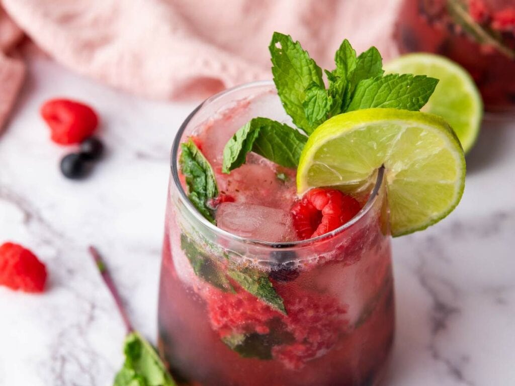 A glass of beverage with ice, fresh mint leaves, raspberries, and lime slices on a marble surface.