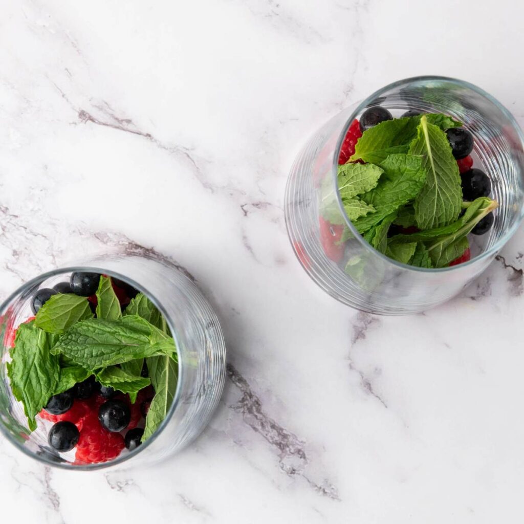 Two glasses on a marble surface, each containing raspberries, blueberries, and fresh mint leaves.