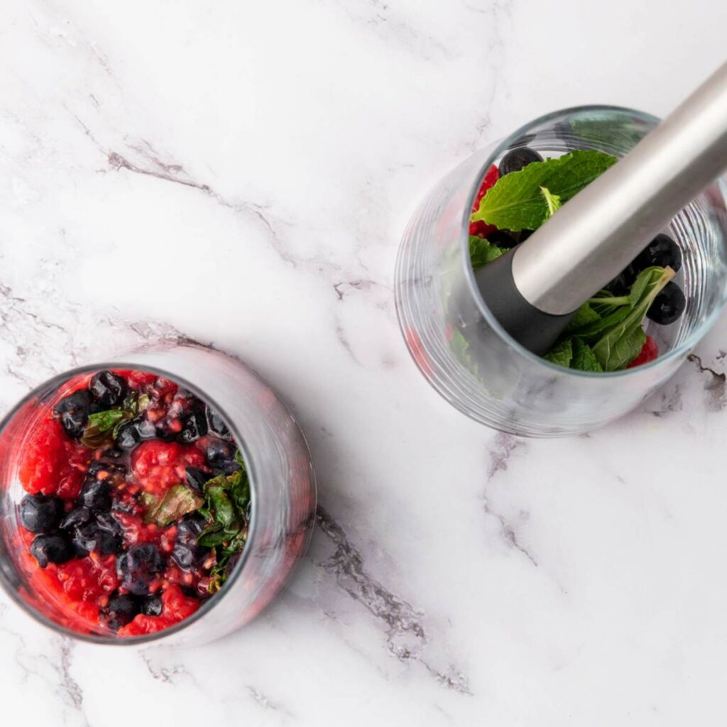 Two glasses on a marble surface, one with crushed berries and mint, the other with whole berries, mint, and a muddler inside.