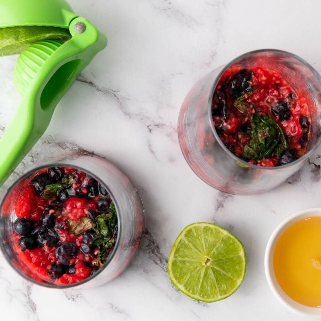 Two glasses with mixed berries and mint, a green citrus squeezer, half a lime, and a small bowl of liquid sweetener.