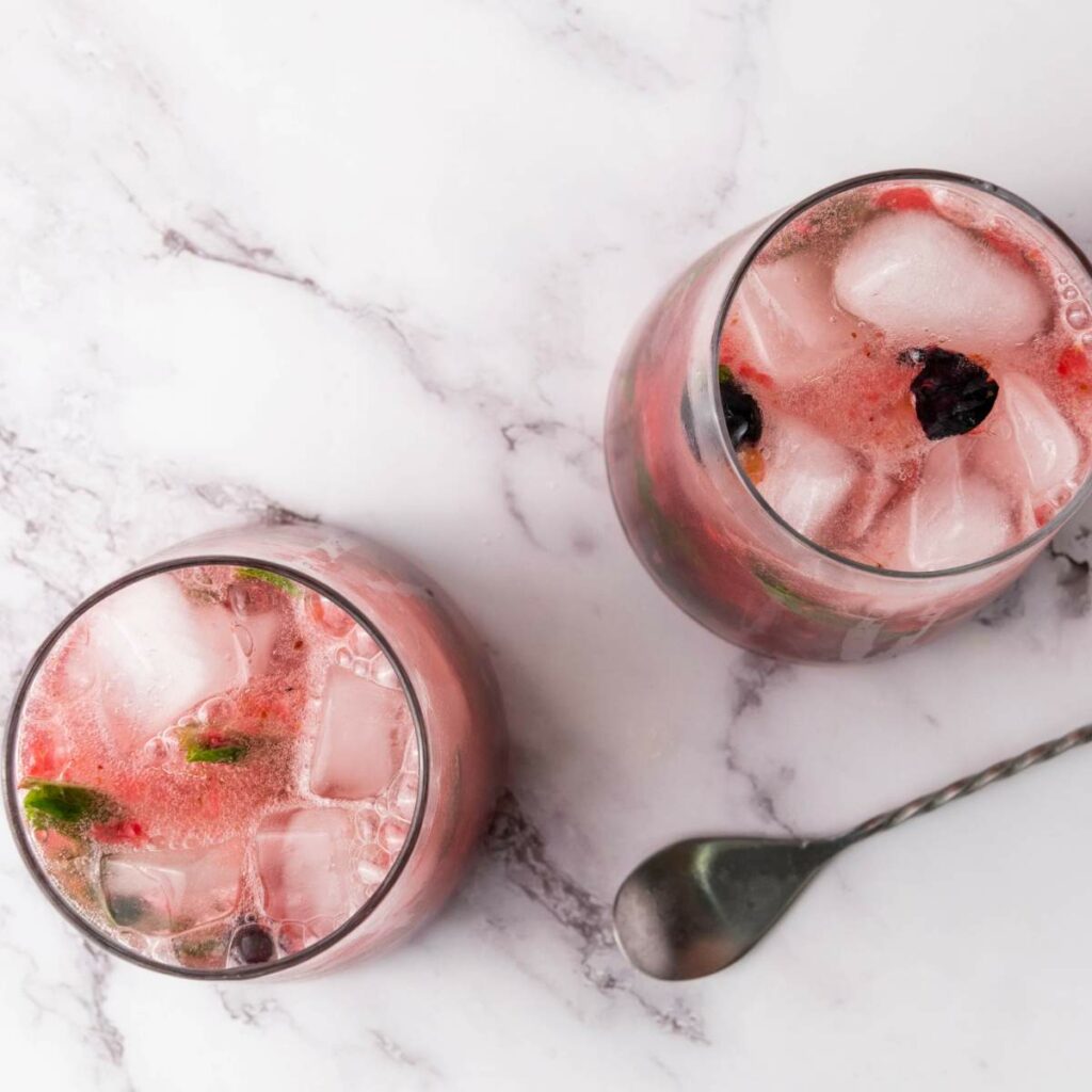Pink berry soda water with ice cubes and mint leaves on a marble surface, next to a metal spoon.