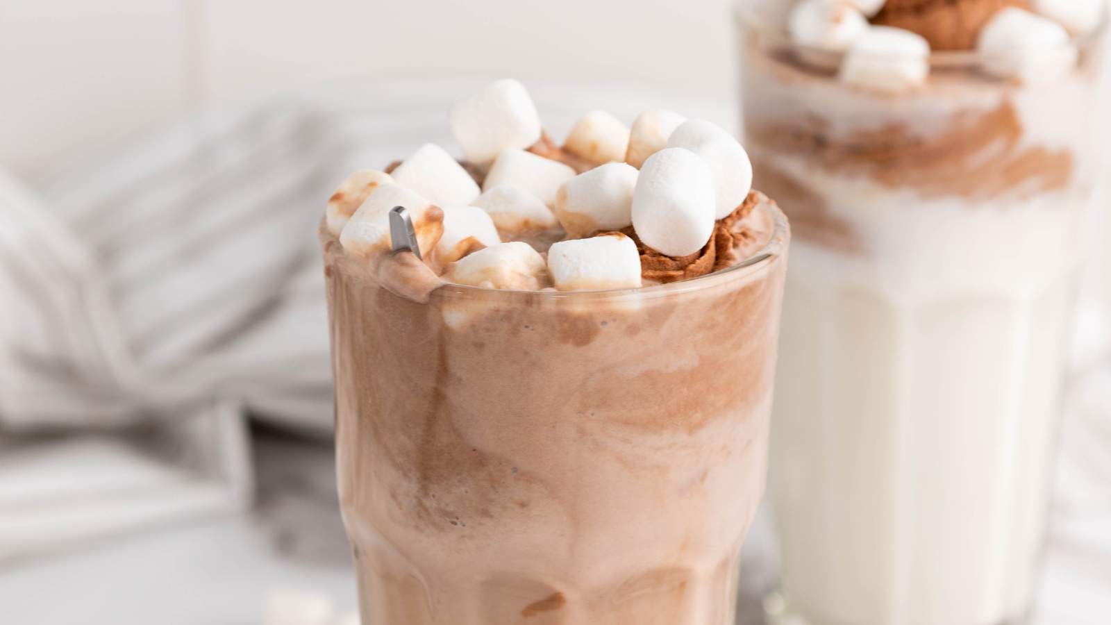 A creamy chocolate drink served in a tall glass. Another glass with a similar drink is partially visible in the background.