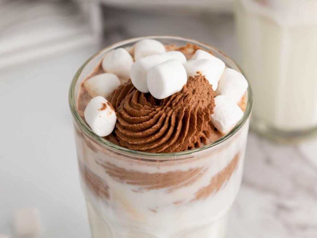 A glass filled with milk topped with swirls of chocolate cream and marshmallows.