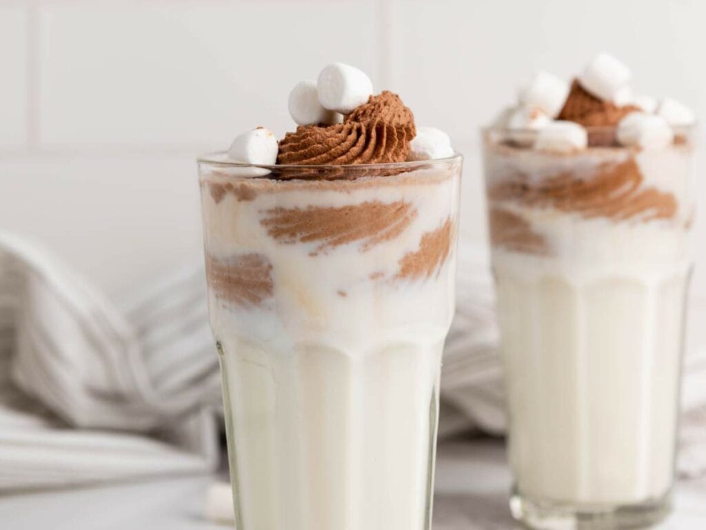 Two glasses of milk with chocolate cream garnished with mini marshmallows next to a white striped cloth on the background.