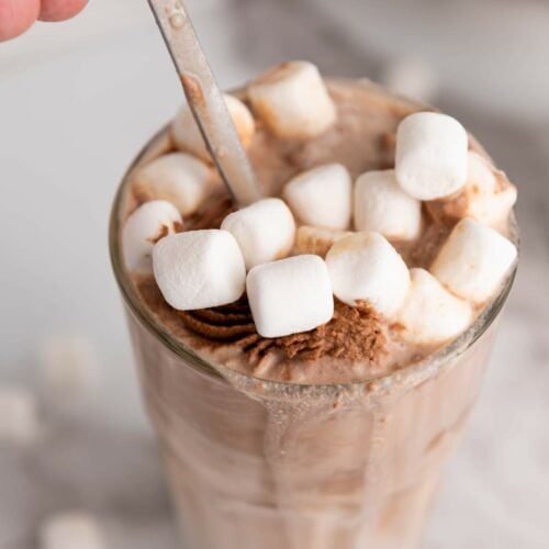 A hand holding the spoon of a glass filled with chocolate milkshake topped with small white marshmallows.