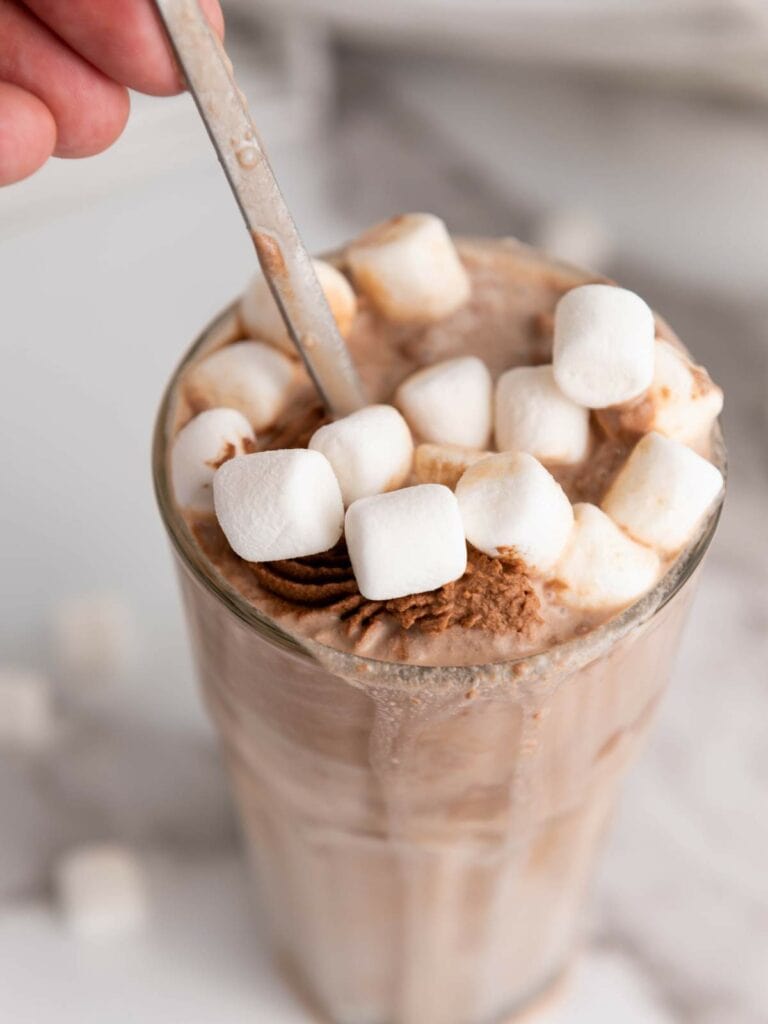 A hand holding the spoon of a glass filled with chocolate milkshake topped with small white marshmallows.