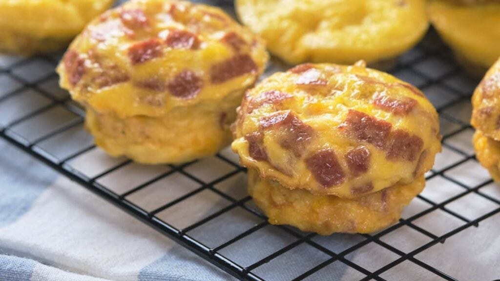 Cheesy egg bites are packed with chunks of ham and baked until golden, are cooling on a wire rack.
