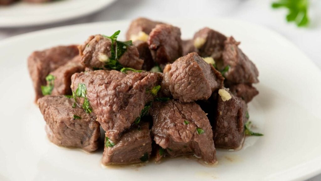 Juicy steak bites served on a white plate garnished with fresh parsley and garlic on a white plate.