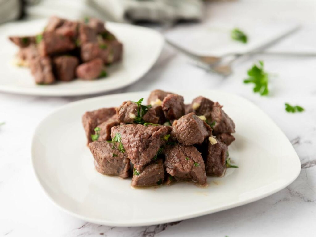 Two plates of steak, garnished with fresh cilantro and garlic on a white marble surface.