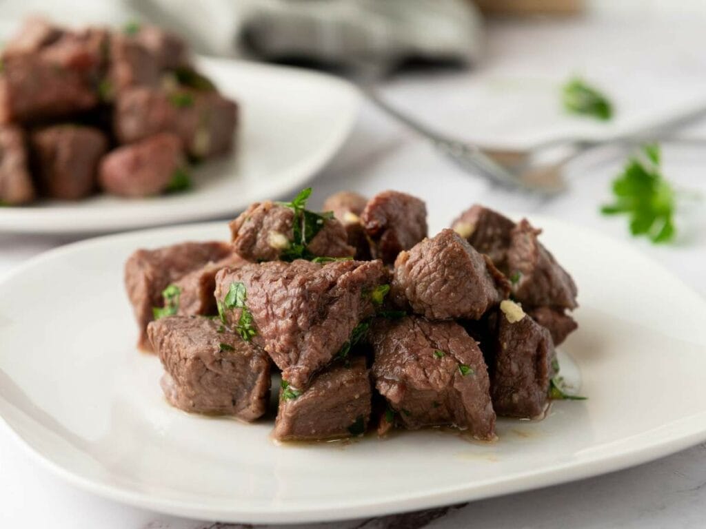Juicy cooked steak served on a white plate with scattered parsley and a second plate in the background.
