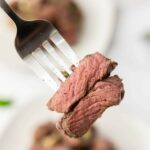 A fork holding pieces of cooked steak, with visible spices and two plates on the background.