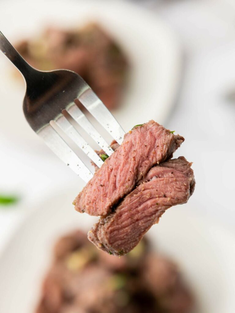 A fork holding pieces of cooked steak, with visible spices and two plates on the background.