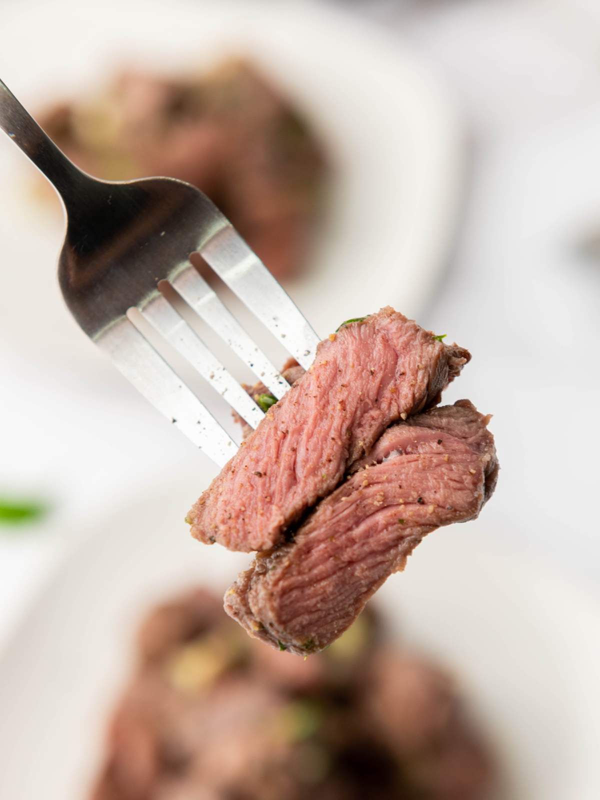 A fork holding pieces of cooked steak, with visible spices and two plates on the background.