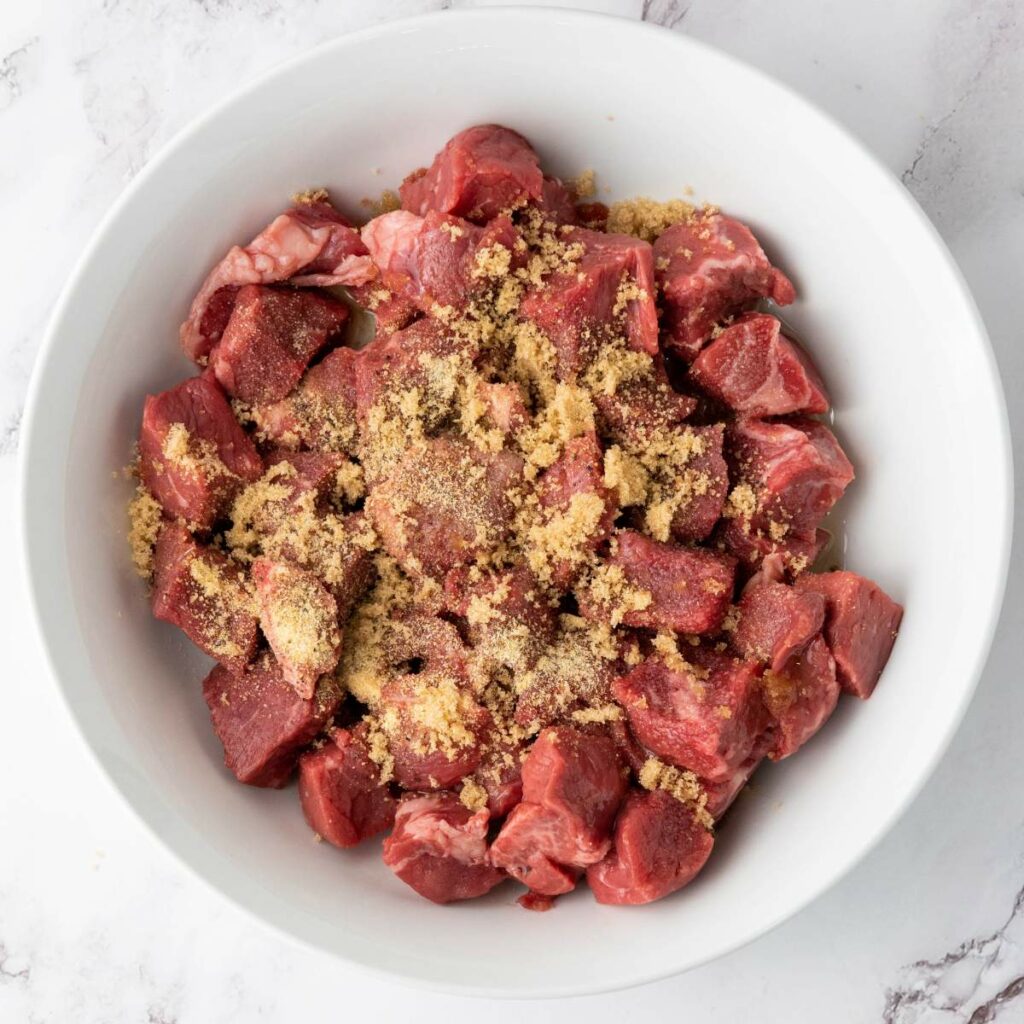 Cubed steak pieces in a bowl are topped with seasonings, ready to be mixed.
