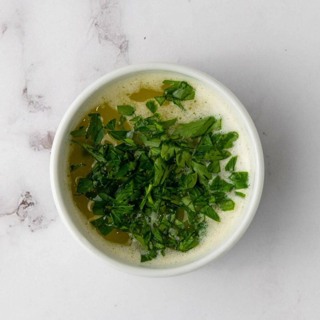 Butter, garlic, and parsley are combined in a small bowl on a marble surface.