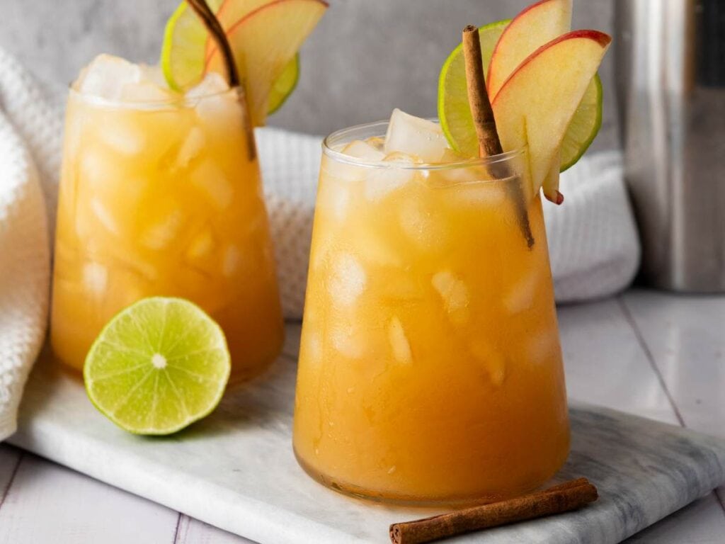 Two glasses of orange iced drink garnished with apple slices, lime wheels, and cinnamon sticks sit on a marble board.