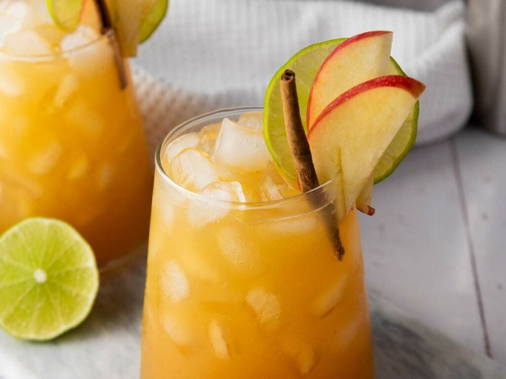 A glass of orange-hued iced drink garnished with lime and apple slices, a cinnamon stick, and ice cubes.