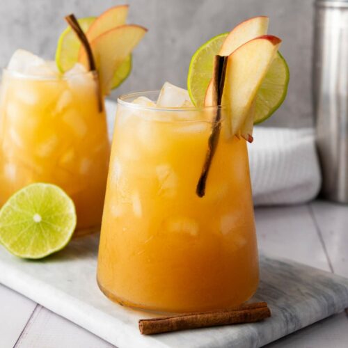 A glass of iced orange cocktail garnished with apple slices, a lime wheel, and a cinnamon stick, placed on a marble board.