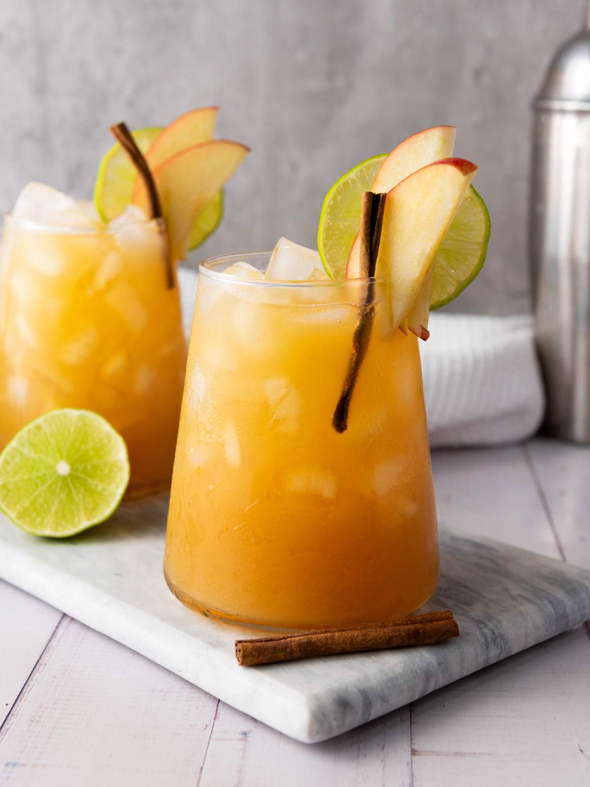 A glass of iced orange cocktail garnished with apple slices, a lime wheel, and a cinnamon stick, placed on a marble board.