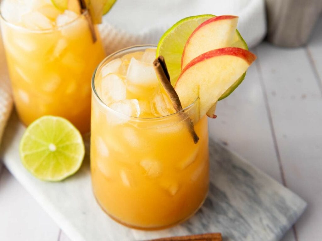A glass of orange-colored iced drink garnished with lime and apple slices and a cinnamon stick, with ice cubes and a halved lime nearby.