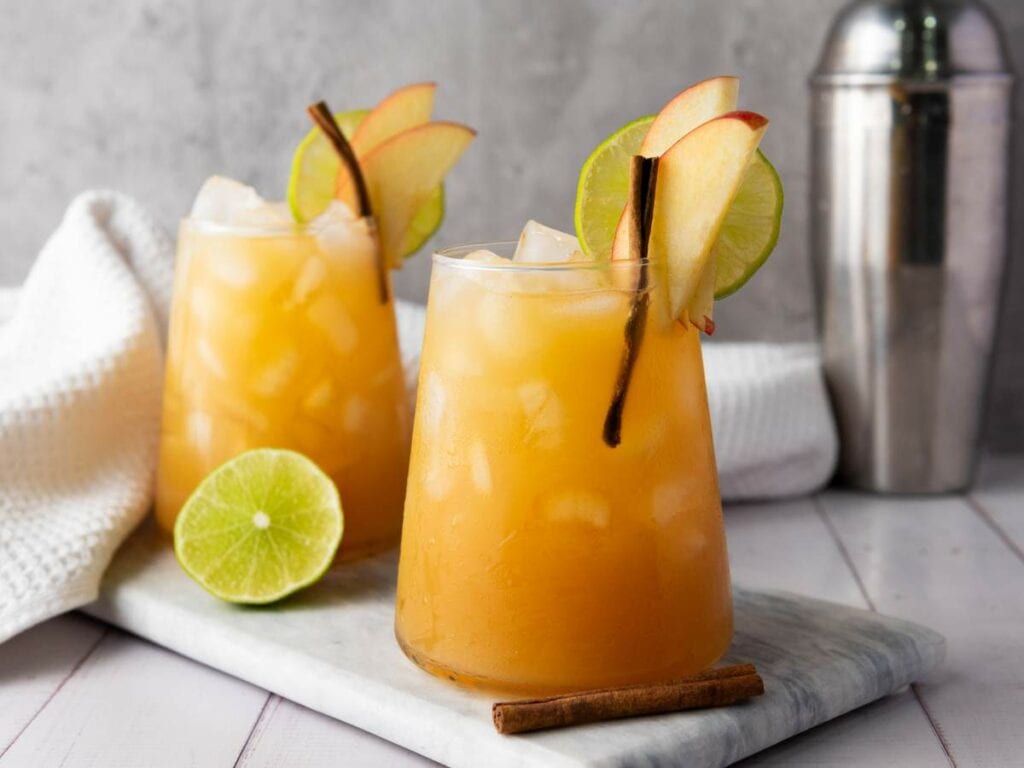 Two glasses of iced orange beverage on a marble tray with a lime half, cinnamon stick, white towel, and cocktail shaker in the background.