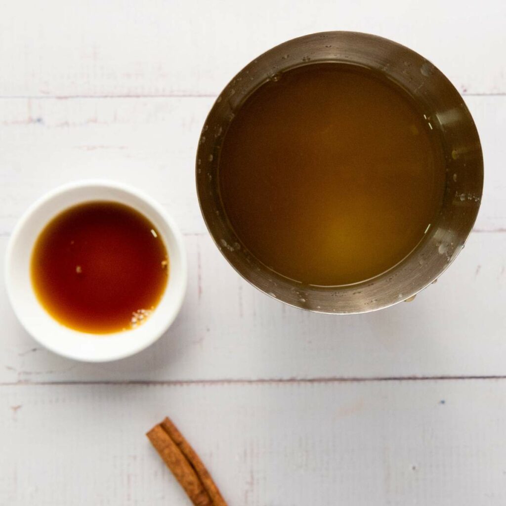 A metal shaker with a yellowish liquid, a small white bowl with a brown liquid, and a cinnamon stick.