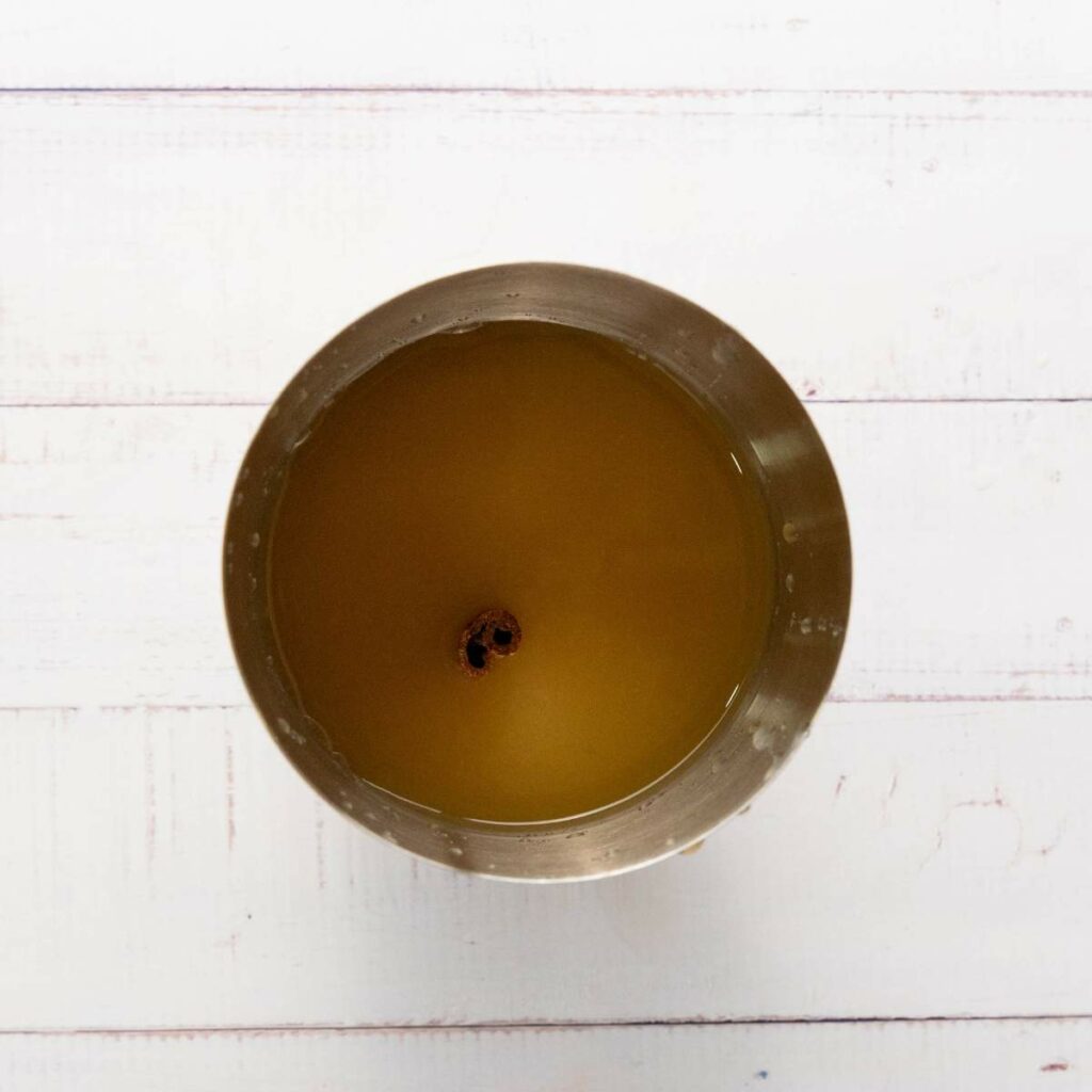 A metal shaker filled with yellowish-brown liquid and a cinnamon stick floating in the center.
