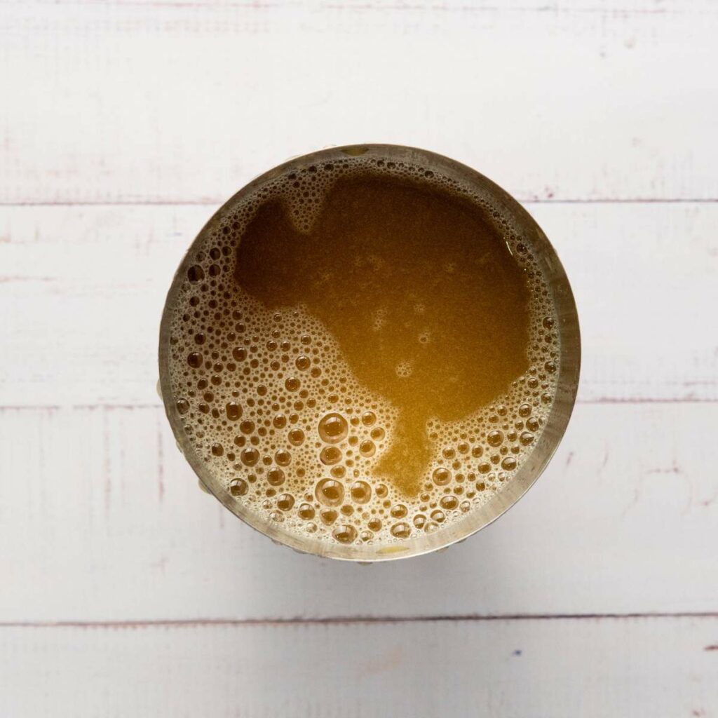 A top-down view of a metal shaker filled with frothy brown liquid on a white wooden surface.