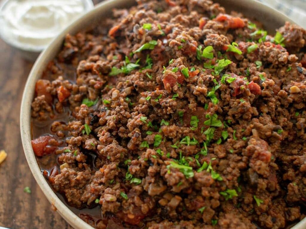 A bowl of cooked ground beef with tomatoes and herbs, garnished with chopped parsley, with a side of sour cream in the background.